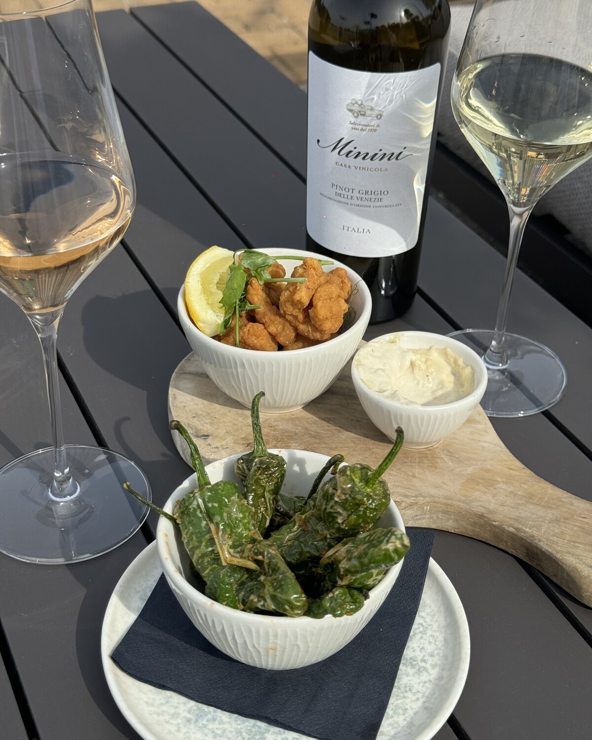 A bottle of chilled white wine with two glasses, padrón peppers and crispy nibbles on a sunlit table in the Courtyard at The Glasshouse, Windlesham